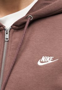 Bruine Nike hoodie met ritssluiting, trekkoord en wit geborduurd logo op de borst, gedragen over een wit shirt.
