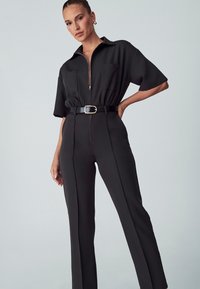 Zwarte jumpsuit met korte mouwen, ritssluiting, twee borstzakken, een ceintuur in de taille en rechte pijpen. Glad weefsel met een aansluitende uitstraling.