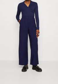 Mörkblå långärmad omslagstopp i kombination med högmidjade vida byxor med en slät textur och kontrasterande kraftiga svarta skor.