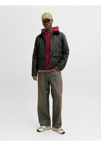 Veste noire cirée portée sur un sweat à capuche bordeaux, associée à un jean large gris clair et des baskets beige. Casquette olive avec détail texte.