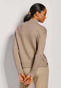 Beige gestrickter Pullover mit hohem Kragen, geripptem Saum und seitlichen Knöpfen; kombiniert mit passenden Hosen. Glatte Textur, enganliegendes Design.