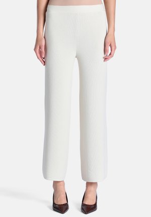 Pantalon blanc à jambes larges porté par une personne debout, associé à des talons hauts foncés à bout pointu, sur un fond uni.