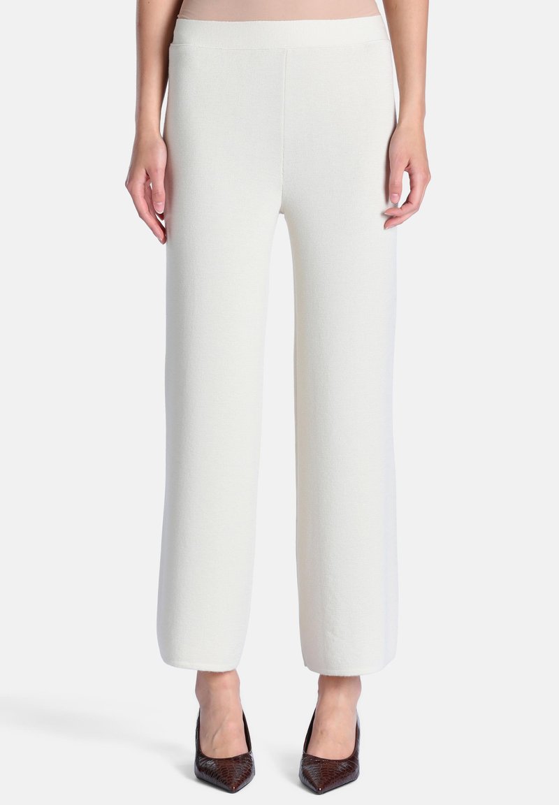 Pantalon blanc à jambes larges porté par une personne debout, associé à des talons hauts foncés à bout pointu, sur un fond uni.