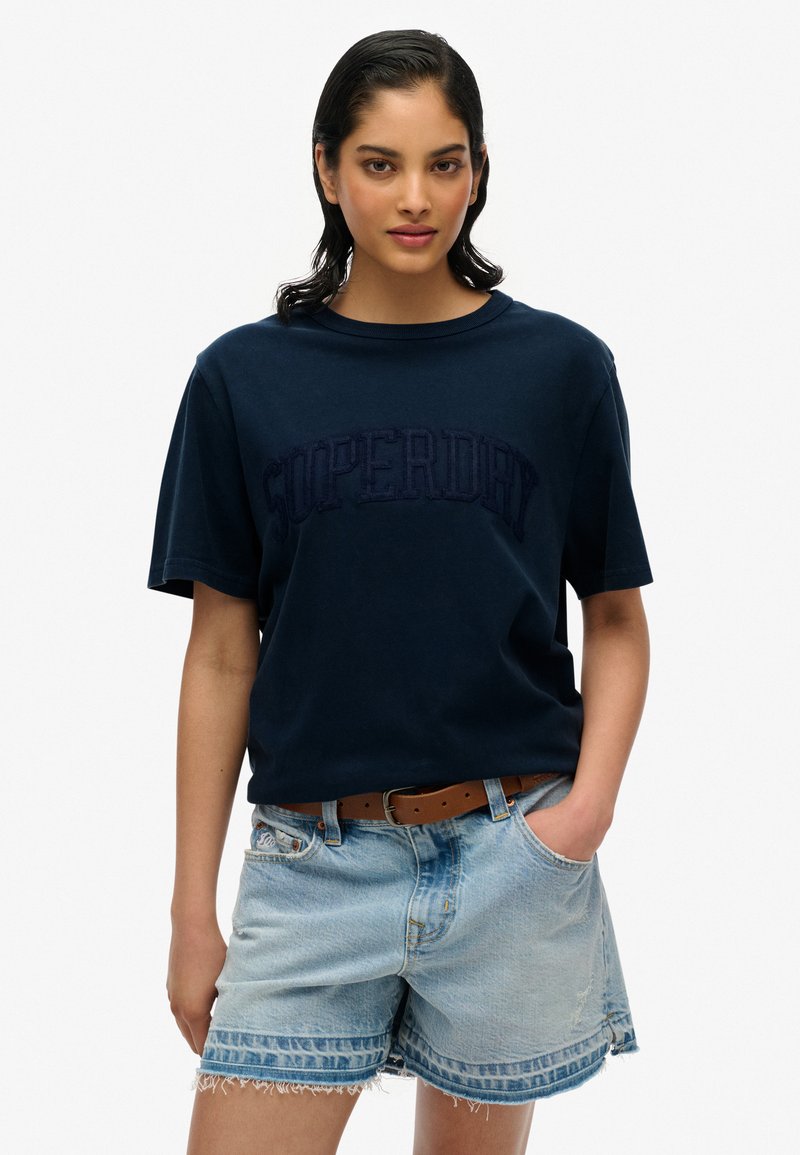 Marineblå bomulds T-shirt med korte ærmer og 'Superdry' logo i et hævet design, parret med lyse denimshorts og et brunt bælte.