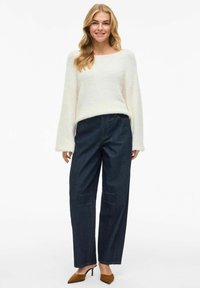 White, textured sweater with wide sleeves paired with dark blue, high-waisted jeans featuring front pockets and light stitching. Brown heels.