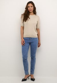 Pull beige à manches courtes avec un col rond, associé à un jean skinny bleu et des escarpins noirs à bouts pointus avec des embellissements.
