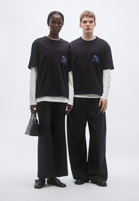 Čierne tričká s detailom vrecka, pod nimi dlhé biele rukávy. Voľné čierne nohavice. Modelka drží malú čiernu kabelku.