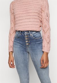 Suéter de punto rosa claro con rayas texturizadas y mangas caladas, combinado con jeans de mezclilla azul de cintura alta con cinco botones.