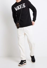 Persona che indossa un felpa nera Vans, pantaloni bianchi, sneakers in bianco e nero e una piccola borsa a tracolla con stampa leopardata.