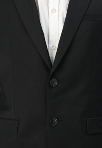 Blazer noir avec deux boutons noirs, poche poitrine simple et col cranté. En dessous, une chemise blanche à boutons est visible.