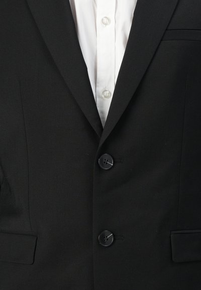 Blazer noir avec deux boutons noirs, poche poitrine simple et col cranté. En dessous, une chemise blanche à boutons est visible.