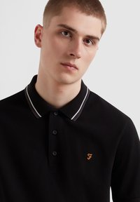 Polo noir avec une patte de boutonnage, doté d'un col rayé contrastant et d'un logo brodé orange sur la poitrine.