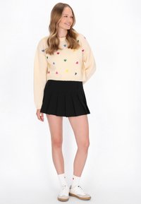 Crème gebreide trui met meerkleurige hartpatronen, gecombineerd met een zwarte geplooide minirok. Het model draagt witte sokken en sneakers.