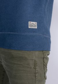 Sudadera de algodón azul con dobladillo acanalado, que presenta una etiqueta con detalles de la marca. Combinada con pantalones verde oliva, que exhiben un acabado texturizado.