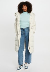 Donna con capelli ricci che indossa un lungo cappotto bianco imbottito, un dolcevita azzurro chiaro, jeans blu e sneakers blu e bianche, in piedi contro uno sfondo bianco.