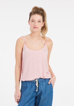 Joven mujer con cabello rubio recogido en una coleta, vistiendo una camiseta sin mangas rosa claro y pantalones azules con cordón, de pie frente a un fondo blanco.