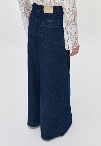 Γυναίκα που φοράει σκούρο μπλε τζιν παντελόνι φαρδιάς γραμμής με ψηλή μέση και πίσω τσέπες, σε συνδυασμό με λευκό μακρυμάνικο τοπ από διάφανη δαντέλα με λουλούδια.