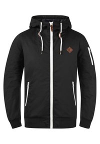 Veste noire avec capuche, fermeture éclair à l'avant, détails de fermeture éclair blancs, et deux poches latérales. Comprend un patch logo en cuir marron sur la poitrine.