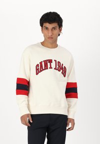 Kremowa bluza z logo "GANT 1949". Posiada wyraźne czerwone i granatowe paski na rękawach. Wykonana z miękkiego materiału, z okrągłym dekoltem i ściągaczami na rękawach.
