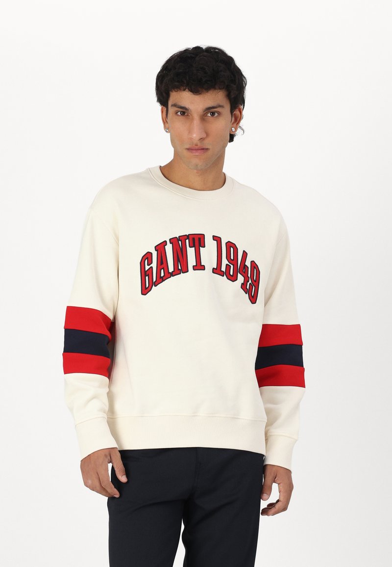 Kremowa bluza z logo "GANT 1949". Posiada wyraźne czerwone i granatowe paski na rękawach. Wykonana z miękkiego materiału, z okrągłym dekoltem i ściągaczami na rękawach.