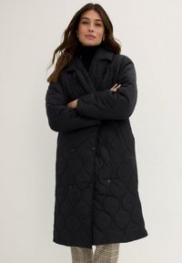 Cappotto imbottito nero con design doppiopetto, ampio colletto e sottile motivo ricamato. Abbinato a un top a collo alto e pantaloni con motivo.
