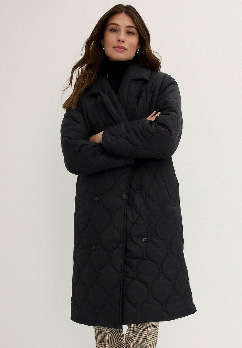 Cappotto imbottito nero con design doppiopetto, ampio colletto e sottile motivo ricamato. Abbinato a un top a collo alto e pantaloni con motivo.