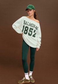 Felpa oversize color crema con "BALMORAL 1852" in verde scuro, abbinata a leggings verde scuro, sneakers bianchi e verdi, e cappellino verde.
