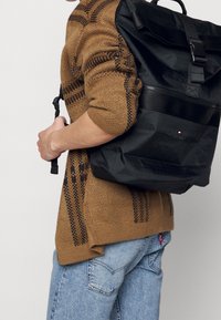 Black roll-top backpack with textured fabric, adjustable straps, and a metallic buckle. The woven brown sweater has a geometric pattern.
