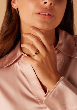 Gouden ring met een driehoekige zwarte steen, gedragen aan een hand die op de kin rust; model in een zachtroze satijnen shirt.