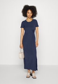 Granatowa sukienka maxi z okrągłym dekoltem, krótkimi rękawami i detalem w postaci węzła z boku. Gładki materiał i rozcięcie po boku. Połączona z jasną torebką.