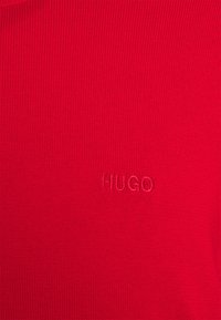 Tissu tricoté rouge avec un logo "HUGO" brodé subtilement en fil rouge assorti, centré sur le matériau.