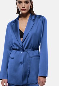 Blazer blu in satin con vita cinturata, colletto a revers e chiusura con bottoni. Presenta tasche laterali e una texture liscia.