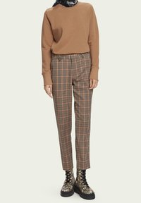 Bruine sweater met lange mouwen, geruite broek in oranje, zwart en crème, gecombineerd met zwart patroonlaarzen. Effen achtergrond.