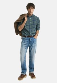 Mann in einem grün-blau karierten Hemd, hellblauen Jeans, braunem Gürtel und braunen Schuhen, der eine braune Jacke über der Schulter hält.