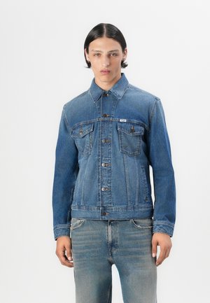 Un mannequin portant une veste en denim sur un t-shirt clair et un jean déchiré, se tenant devant un fond blanc uni.