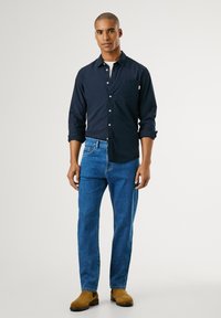 Tengerészkék gombos ing, mellzsebbel, kék farmernadrággal és barnás bokacsizmával párosítva. Az ing feltekert ujjakkal és laza szabással rendelkezik.