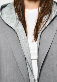 Jasnoszara kurtka o fakturowanym wykończeniu, rozpięta, odsłaniająca miękki szary kaptur bluzy pod spodem oraz białą koszulkę.