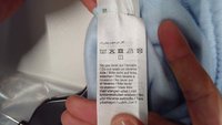 Textílápolási címke fehér szövet háttéren, több nyelven, mosási szimbólumokkal és kék textil mintával.