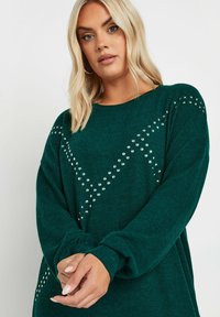 Maglione verde smeraldo con maniche lunghe, caratterizzato da decorazioni in argento a forma di borchie in un motivo zigzag sulla parte anteriore. Tessuto morbido e strutturato.