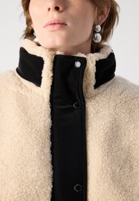 Beige nep-bont jas met een zwarte, glanzende kraag. Voorzien van een drukknoopsluiting en zichtbare textuur op de vacht. Eenvoudig en functioneel ontwerp.
