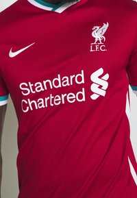 Camiseta de fútbol roja con diseño de cuello en V, logo de Nike en blanco y "Standard Chartered" impreso en letras blancas en negrita. Acentos verdes en las mangas.