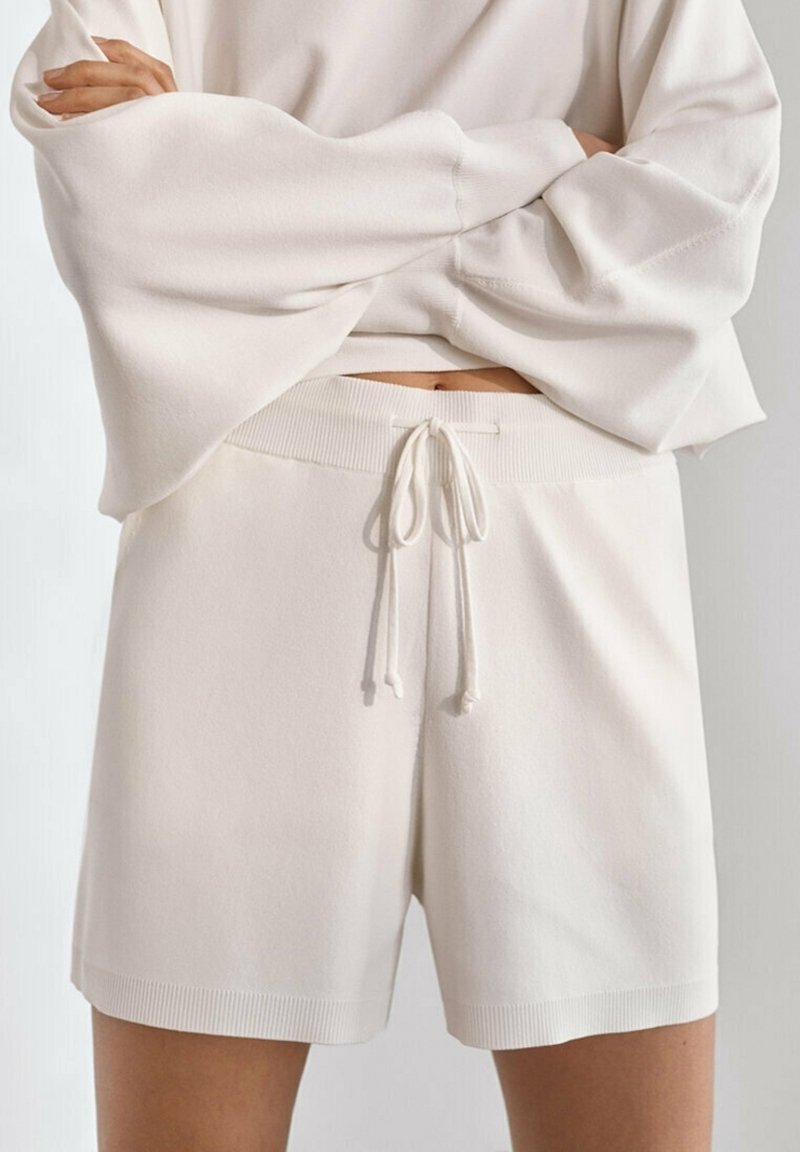 Personne portant un short ample en tricot blanc cassé avec cordon de serrage et un haut assorti à manches longues larges, les bras croisés sur la poitrine.