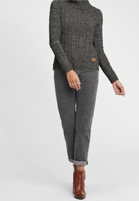 Personne portant un pull en tricot torsadé gris foncé, un jean gris retroussé et des bottines marron, debout les bras détendus.