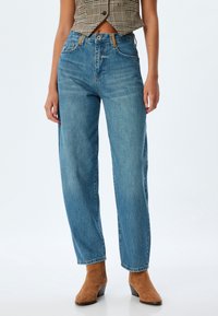 Jeans de mezclilla de talle alto en azul claro con un corte holgado, que presentan un cierre de botones, trabillas para cinturón y piernas rectas, combinados con botas de tacón marrones.