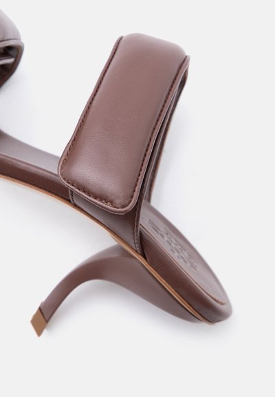 Καφέ δερμάτινο πέδιλο με τακούνι, με φαρδιά επενδεδυμένη λουρίδα πάνω από το πόδι και καμπυλωτό τακούνι στιλέτο σε λευκό φόντο.