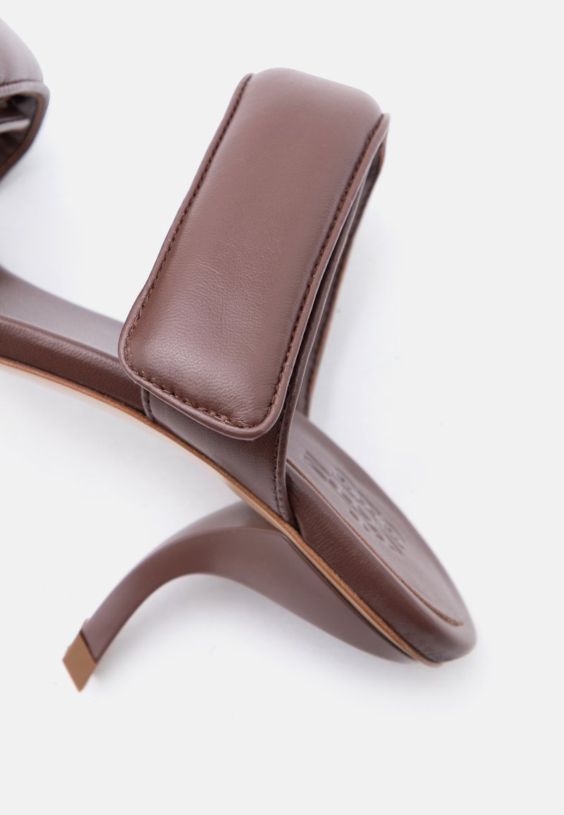 Bruine leren sandaal met hak, voorzien van een brede, gewatteerde band over de voet en een gebogen stilettohak, op een witte achtergrond.