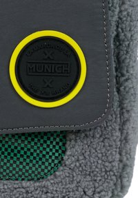 Bolso de tela gris con un parche de logo negro y amarillo, textura suave y un acento de tablero de ajedrez verde. Bordes cosidos en toda la superficie.