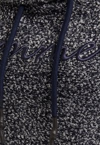 Neulepusero laivastonsinisessä ja harmaassa teksturoidulla kankaalla, jossa on mustapäiset kiristysnyörit ja etuosassa kirjailtu teksti.