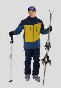 Ski jakke i blå og gul, med lynlåslommer. Sorte handsker. Holder på ski og stave. Grå skistøvler. Solid design, funktionelle egenskaber.