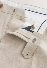 Pantaloni beige in lino parzialmente sbottonati, che mostrano una fodera interna bianca con rifinitura blu navy, un bottone e chiusura a gancio metallico in vita.